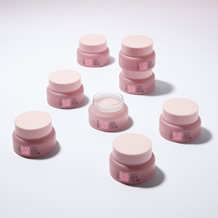 Zdjęcie na różowym tle: po środku otwarty różowy słoiczek "Kimoco Glass Skin Luminous Aqua Cream" z widocznym kremem na wierzchu, z rozmytymi słoiczkami w tle po obu stronach i odbiciem na lustrzanej powierzchni.